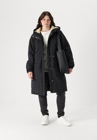 Manteau parka noir avec une capuche en fausse fourrure légère, des poignets ajustables et des poches avant. Porté avec un pantalon gris et des chaussures blanches. Design simple.