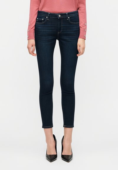 Σκούρα μπλε στενά τζιν από denim, με πέντε τσέπες, διακριτικό γαζί, και μέση μεσαίου ύψους, συνδυασμένα με μαύρες γόβες.