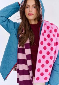 Modrá prešívaná bunda s kvitnúcou vnútornou podšívkou, v páre s ružovým šálom s veľkými bodkami a fialovým pruhovaným akcentom.