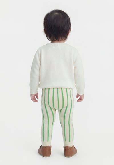 Παιδί σε όρθια στάση με την πλάτη προς την κάμερα, φοράει άσπρο πουλόβερ, κρεμ κολάν με πράσινες κάθετες ρίγες και καφέ παπούτσια, σε λευκό φόντο.