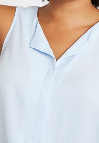 Top senza maniche di colore azzurro chiaro realizzato in un tessuto morbido, dotato di colletto a punta e chiusura frontale con bottoni, con una texture liscia e dettagli minimi.