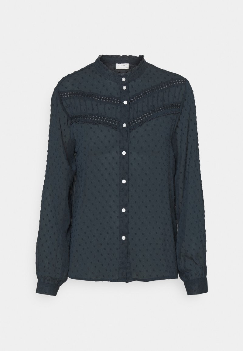 Dark blue blouse made of textured fabric with a dotted pattern. Features a round collar, button closure, and ruffled detailing across the chest.