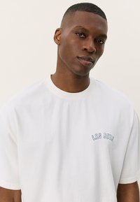 Tricou alb din bumbac, cu decolteu rotund, mâneci scurte și text brodat albastru "LES DEUX" pe partea stângă a pieptului. Textură netedă.