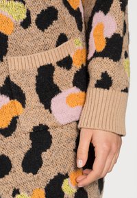 Sweter z dzianiny w beżowym kolorze z kolorowymi łatami w panterkę w czarnym, różowym, pomarańczowym i zielonym kolorze. Z mankietem prążkowanym i detalem kieszeni.