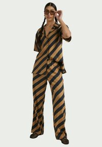 Γυναίκα με πλεξούδα που φοράει oversized καφέ και μαύρο πουκάμισο με διαγώνιες ρίγες και παντελόνι, ρυθμίζοντας μεγάλα τετράγωνα γυαλιά ηλίου.