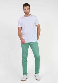 Witte T-shirt met korte mouwen en ronde hals, gecombineerd met lichtgroene skinny broek en witte sneakers met bruine zolen. Neutrale achtergrond.