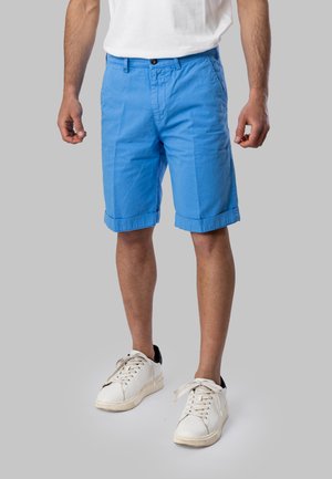 Uomo che indossa pantaloncini azzurri vivaci con risvolto, sneakers bianche e una t-shirt bianca, in piedi davanti a uno sfondo grigio.