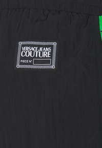 Svart tyg med en Versace Jeans Couture-etikett i vit text och en sydd kant. Inkluderar gröna accenter på sidan.