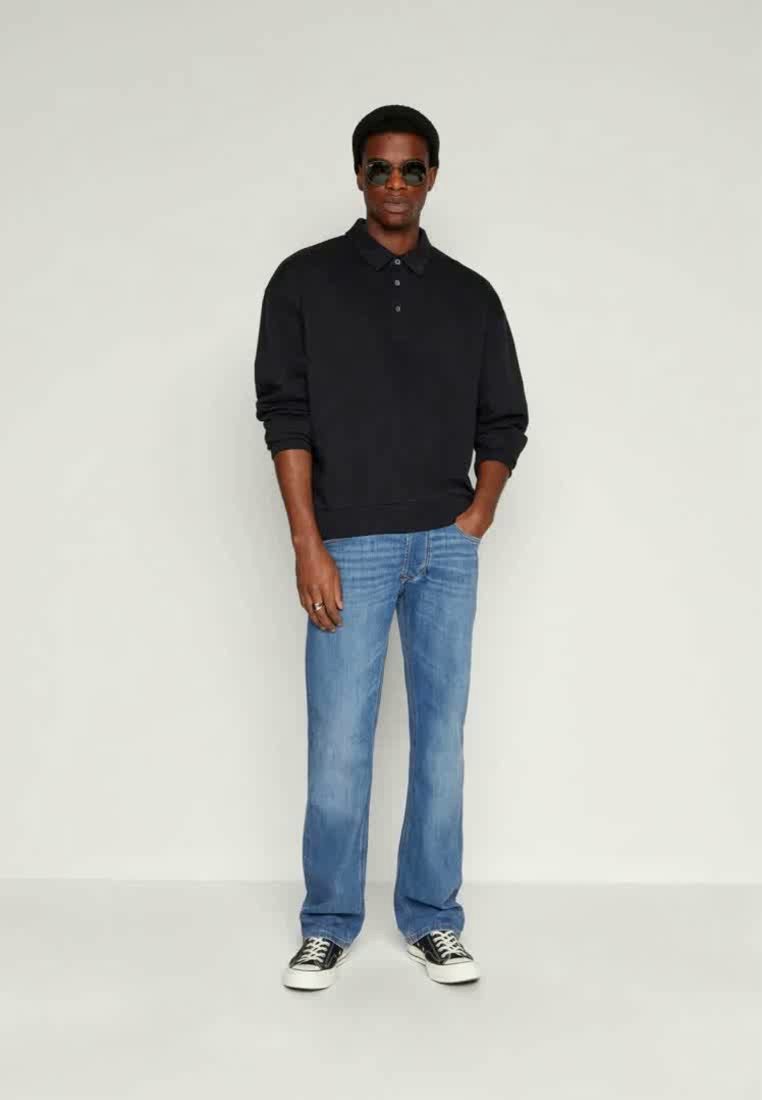 Polo bleu marine, jeans droits bleu délavé et baskets noires. Les accessoires comprennent des lunettes de soleil noires rondes. Pose debout contre un fond neutre.