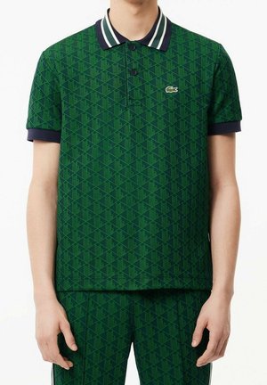 Homme portant un polo à motifs verts avec un col et des poignets de manches bleu marine, associé à un pantalon vert à motifs assorti.