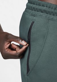 Groene sweatpants met een gestructureerd oppervlak, voorzien van een zwarte ritszak. Een hand wordt getoond die de rits aanpast.