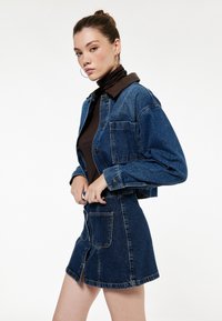 Veste en denim avec une coupe courte, de couleur bleue, fermeture à boutons, et col marron, associée à une jupe mini en denim assortie avec des boutons sur le devant et des poches.