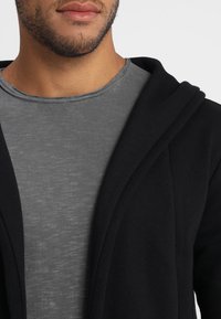 Chaqueta negra con capucha y un acabado texturizado, superpuesta sobre una camiseta gris de cuello redondo. Enfócate en la suave tela y el diseño de abertura frontal.