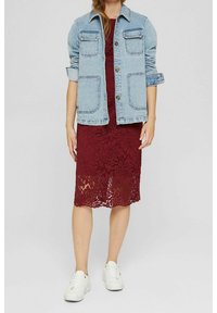 Lichtblauwe denimjack met twee borstzakken over een bordeauxrode kanten jurk. Witte sneakers maken de outfit compleet. De jurk heeft een bloemenkantpatroon.