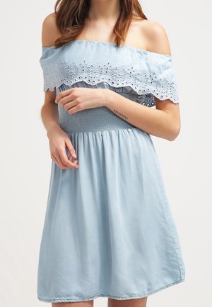 Femme portant une robe bleu clair à épaules dénudées avec bordure en dentelle ajourée et taille froncée, debout avec les mains près de la taille.