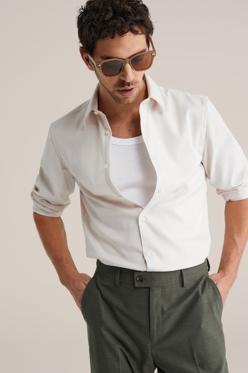 Lysebeige skjorte med knapper over en hvid tanktop, kombineret med mørkegrønne bukser. Brune solbriller fremhæver outfit'et.