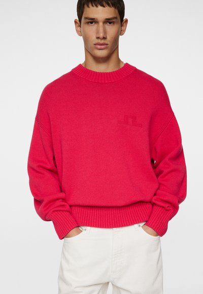 Lyserød strikket sweater med rund hals og ribbede manchetter. Har et diskret logo på brystet, parret med hvide bukser.
