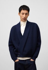 Mörkblå stickad kofta med sjalkrage, textil med struktur och två knappar framtill, bärs över en vit ribbad skjorta.