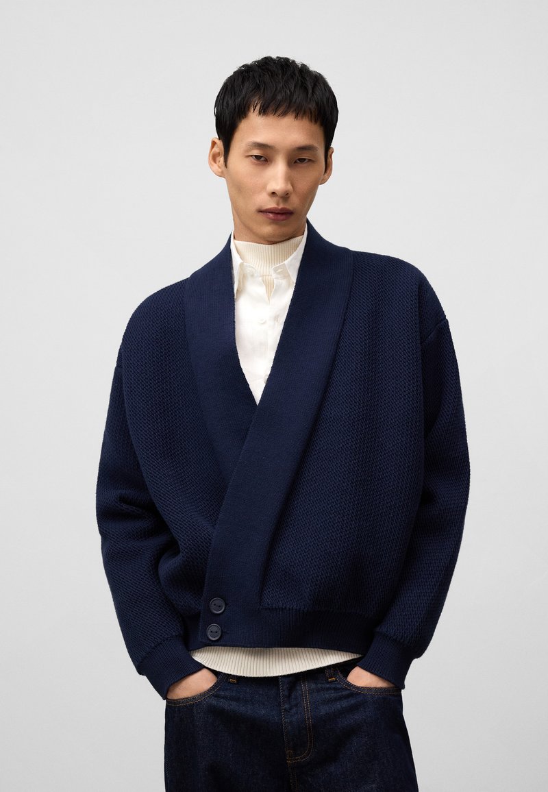 Mörkblå stickad kofta med sjalkrage, textil med struktur och två knappar framtill, bärs över en vit ribbad skjorta.