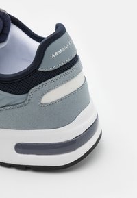 Sapatilhas de camurça azul claro com parte superior em malha, apresentando acabamentos em azul escuro, riscas brancas e uma sola transparente. Logótipo "ARMANI EXCHANGE" no calcanhar.