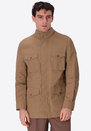 Giovane uomo che indossa una giacca utilitaria color beige con quattro tasche frontali con bottoni, in piedi e che guarda leggermente di lato su uno sfondo uniforme.
