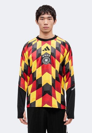 Jeune homme portant un maillot de football à manches longues avec un motif géométrique rouge, jaune et noir et le logo de l’équipe nationale allemande sur la poitrine.