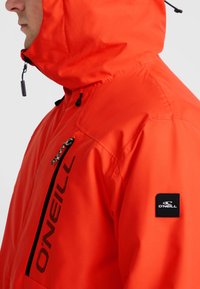 Veste imperméable orange vif avec une poche zippée, capuche réglable, écusson noir et détails de fermeture éclair contrastants noirs.