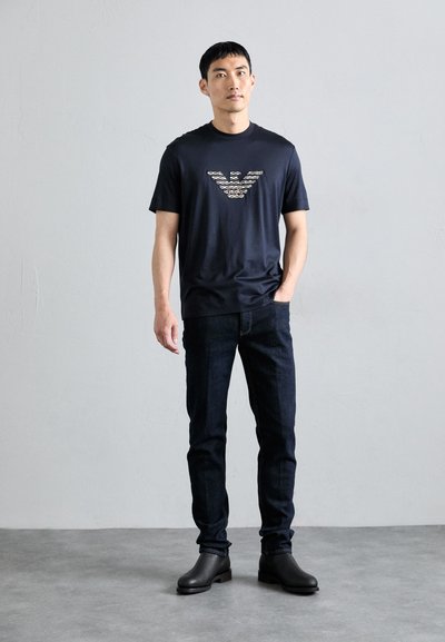 Camiseta de algodón azul marino con un gráfico de logo, combinada con jeans de denim azul oscuro y botas negras hasta el tobillo sobre un fondo gris.
