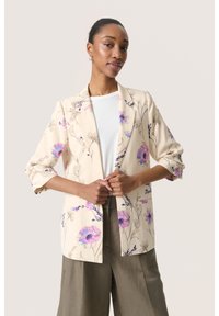 Beige Blazer mit lila Blumenmuster, taillierte Passform, Reverskragen und Dreiviertelärmel, kombiniert mit einem weißen Oberteil und weiten Hosen.