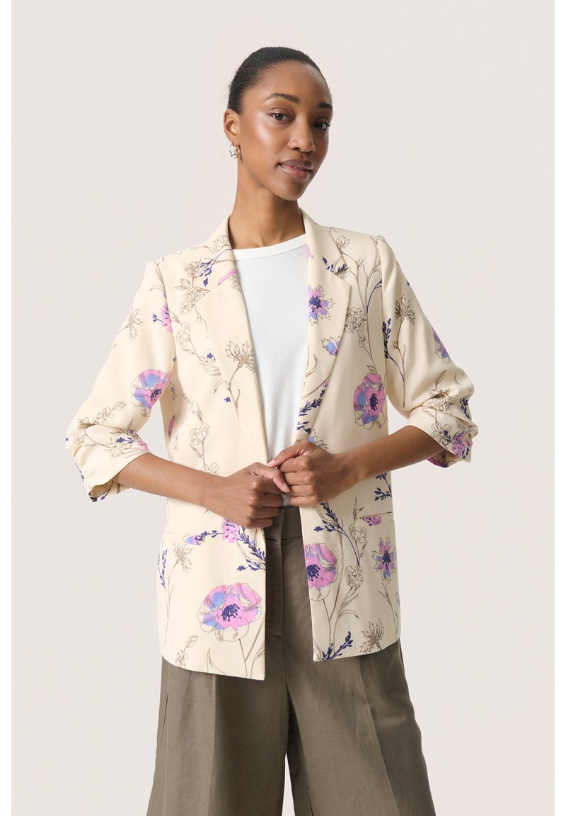 Beige Blazer mit lila Blumenmuster, taillierte Passform, Reverskragen und Dreiviertelärmel, kombiniert mit einem weißen Oberteil und weiten Hosen.