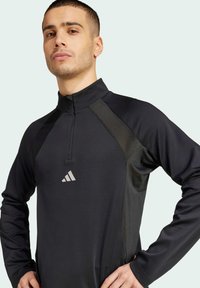 Zwarte sportieve pull met hoge kraag, kwartrits en gestructureerde schouderaccenten. Voorzien van een reflecterend logo in het midden.