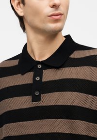 Άνδρας που φοράει πλεκτό πόλο πουκάμισο με μαύρες και καφέ ρίγες, τρία μαύρα κουμπιά, μαύρο γιακά, με ορατό το λαιμό και το κάτω μέρος του προσώπου.