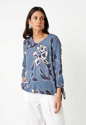 Frau in blau geblümter Langarmbluse mit V-Ausschnitt und weißen Hosen, steht mit den Händen in den Taschen und schaut seitlich vor weißem Hintergrund.