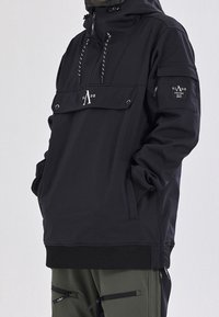Personne portant une veste noire à capuche avec poche avant et écusson sur la manche, mains dans les poches, associée à un pantalon vert olive avec des fermetures éclair.