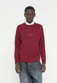 Bordo spalvos medvilninis džemperis su apvalia apykakle, gofruotomis rankogalių juostelėmis ir mažu auksiniu išsiuvinėtu logotipu ant krūtinės.