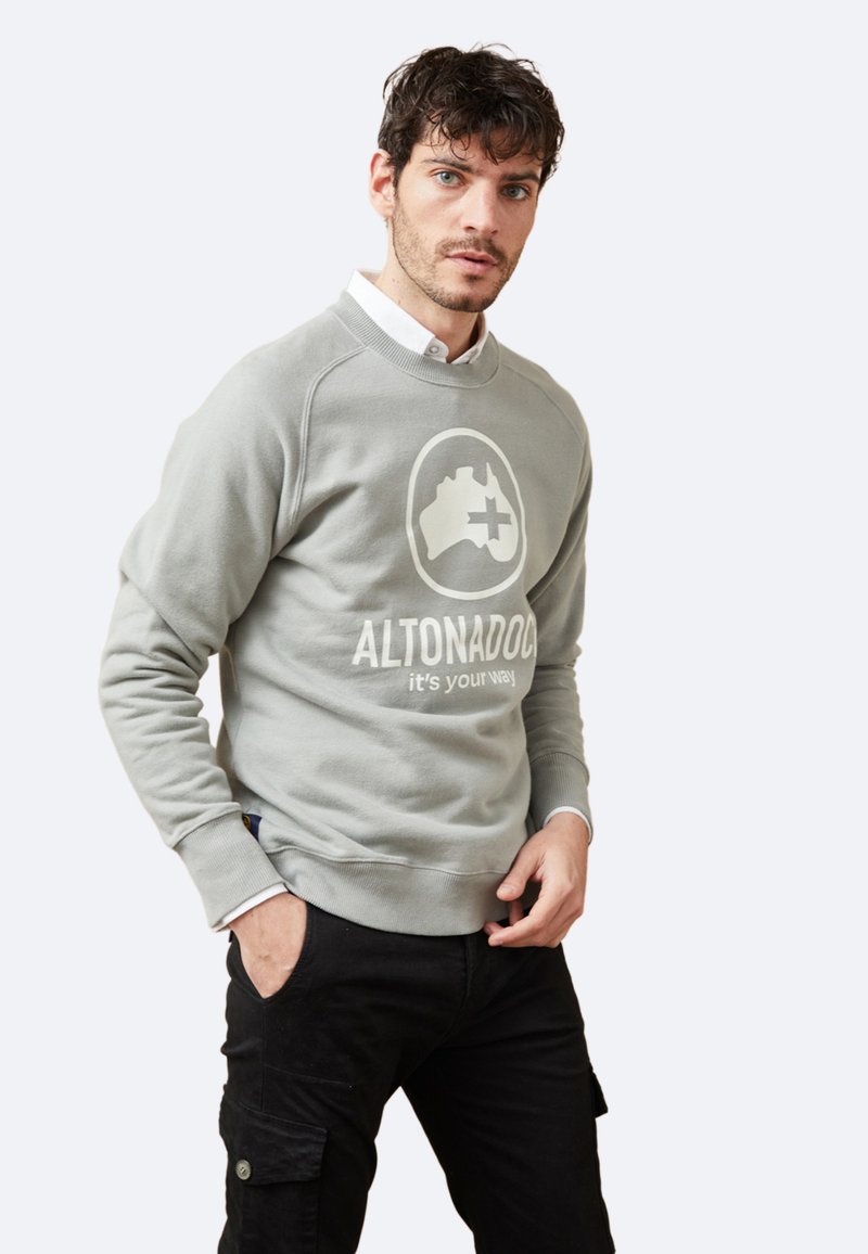 Sudadera gris con cuello redondo, que presenta un gran logotipo y texto en el frente. Combinada con pantalones cargo negros, que muestran bolsillos utilitarios.