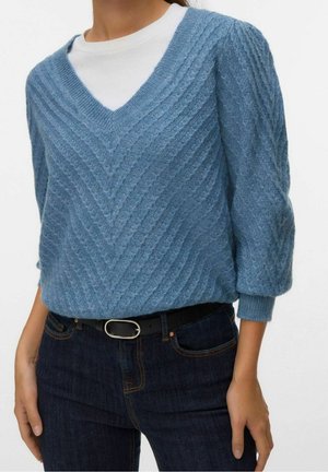 Personne portant un pull bleu à col en V en tricot torsadé sur un top blanc, associé à un jean foncé et une ceinture noire avec une boucle argentée.