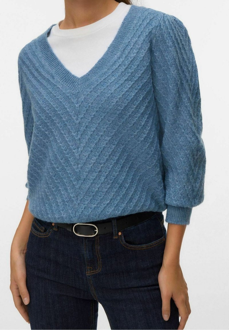Personne portant un pull bleu à col en V en tricot torsadé sur un top blanc, associé à un jean foncé et une ceinture noire avec une boucle argentée.