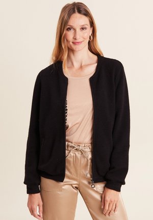 Veste bomber noire texturée avec un col rond, des poignets côtelés et des poches latérales, superposée à un top beige. Tissu doux et coupe décontractée.