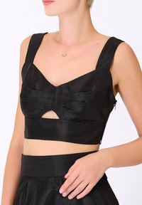 Top court en satin noir avec de larges bretelles, un décolleté en cœur, des détails plissés et une découpe au centre. Fermeture éclair sur le côté.