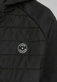 Juodas gobtuvas su siuvinėtu dizainu, lygia tekstūra ir apvaliu logotipo pleistru su užrašu "JACK & JONES" baltomis raidėmis.