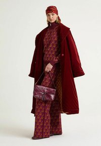 Cappotto di velluto bordeaux, completo fantasia viola e arancione, borsa abbinata, orecchini dorati e sciarpa rossa. Motivi geometrici audaci e strutturati.