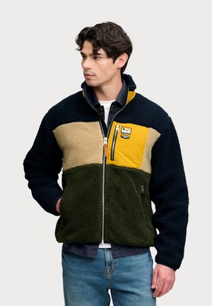 Man wearing a color-block fleece jacket in navy, beige, olive green, and yellow with a front zipper and chest pocket, looking to the side.