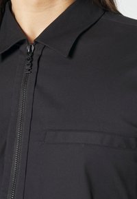 Zwarte blouse met kraag, voorzien van een volledige ritsluiting met een gelabelde ritstrekkker, zachte stofstructuur en een enkele borstzak.