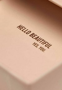 Rosa fyrkantig låda med en slät yta. Insidan har präglad text: "HEJ VACKRA" och "JA. DU." i bruna bokstäver.