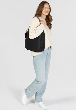Borsa a spalla in pelle nera con design curvo, texture liscia e chiusura con zip. Accentuata da un'etichetta removibile, indossata sulla spalla.
