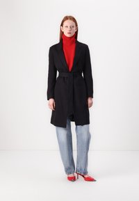 Manteau noir ceinturé porté sur un col roulé rouge, associé à un jean bleu clair et des escarpins rouges à bout pointu avec des accents en argent. Design moderne, texture lisse.