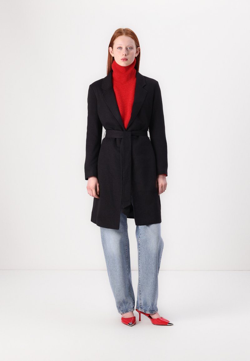 Manteau noir ceinturé porté sur un col roulé rouge, associé à un jean bleu clair et des escarpins rouges à bout pointu avec des accents en argent. Design moderne, texture lisse.