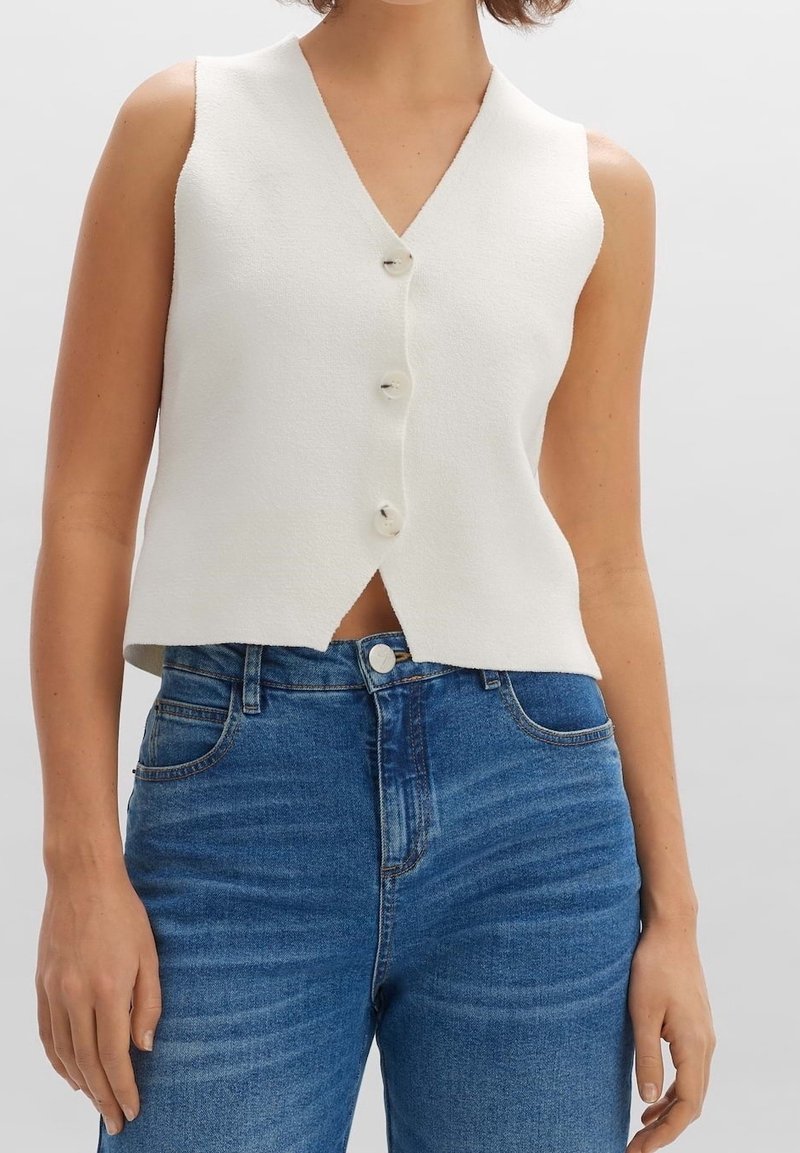 Ärmlös vit skjortväst med V-ringning som är kombinerad med högmidjade blå jeans på en person mot en enfärgad bakgrund.