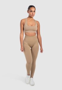 Beige urheilullinen crop-toppi ja korkeavyötäröiset leggingsit, jotka on valmistettu sileästä, joustavasta kankaasta, yhdistettynä valkoisiin urheilukenkiin.